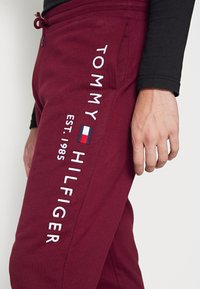 Burgunderfarbene Jogginghose mit weißem Sticktext "TOMMY HILFIGER EST. 1985", ausgestattet mit einem kleinen Flaggenlogo und seitlichen Taschen. Weiches Stoffgefühl.