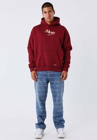 Sudadera burdeos con bolsillo frontal, que presenta un diseño gráfico. Jeans azules con patrón de texto repetido. Zapatillas blancas completan el look.