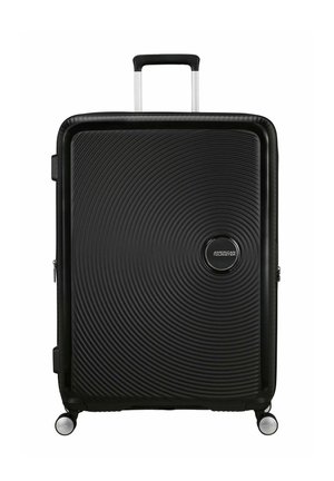 Valise rigide noire avec une texture ondulée, logo argenté, poignée rétractable et quatre roues pivotantes pour une mobilité aisée.