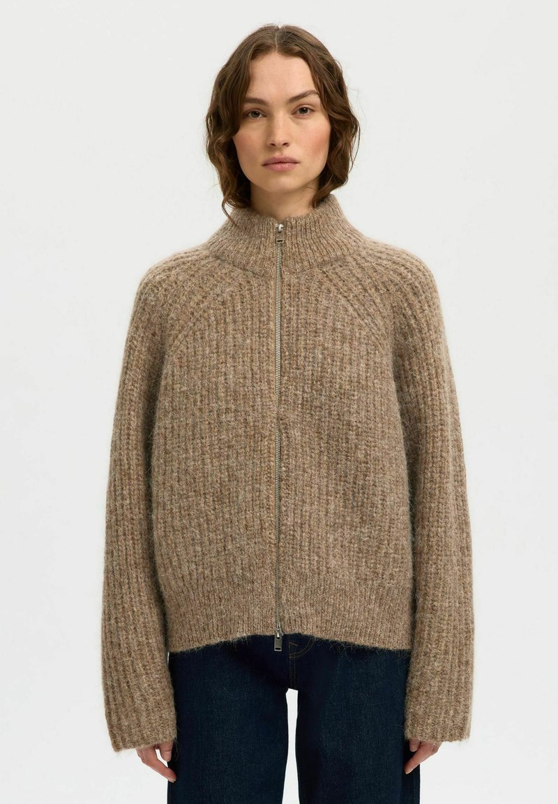 Pull zippé beige avec col montant, texture côtelée et manches oversized, porté avec un jean bleu foncé.