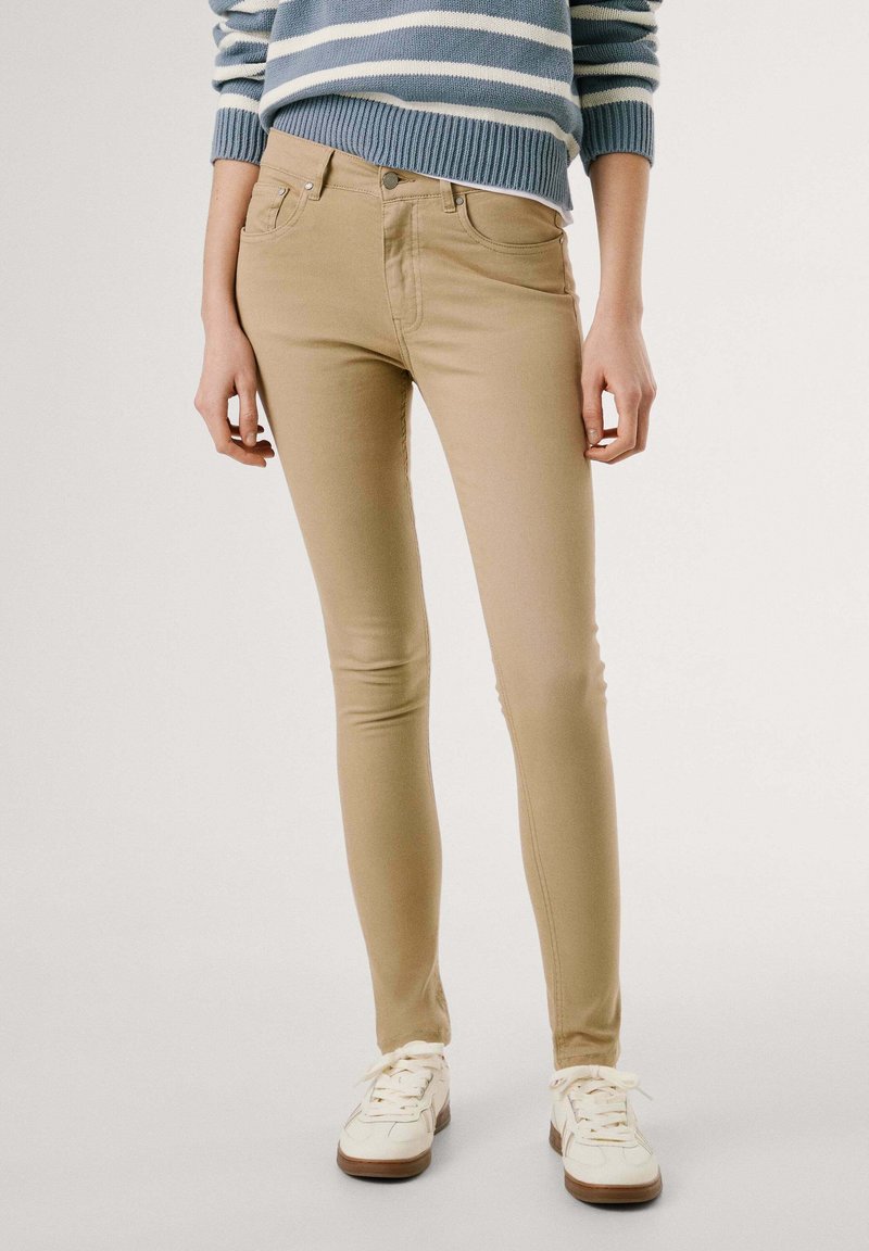 Slim-fite bež jeans iz raztegljive tkanine, z medsešnjo višino pasu, sprednjimi žepi in subtilnimi šivi. V kombinaciji z belimi teniskami.