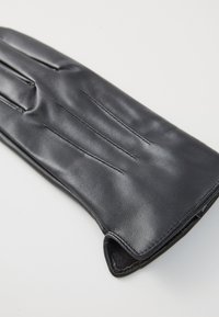 Zwarte leren handschoen met gestikte details, licht gebogen, plat neergelegd op een effen lichte ondergrond, die een zachte textuur en strakke randen laat zien.