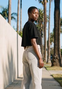 Rövid hajú fiatal fekete rövid felsőt és világos farmert visel, napfényes járdán áll, ahol pálmafák sorakoznak.