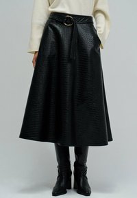 Jupe midi texturée noire avec ceinture, associée à des bottes à talons noires et un pull long à manches crème, se tenant devant un fond uni.