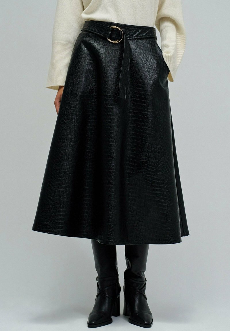 Jupe midi texturée noire avec ceinture, associée à des bottes à talons noires et un pull long à manches crème, se tenant devant un fond uni.