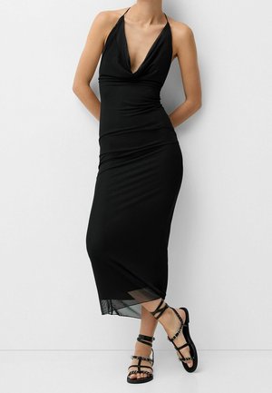 Kobieta ubrana w czarną, bezrękawową sukienkę midi na szyję, nosząca czarne płaskie sandały na paski, stojąca na tle białej ściany.