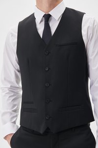 Schwarze Weste aus glattem Stoff, mit sechs Knöpfen, zwei Fronttaschen und einem spitzen Saum. Getragen über einem weißen Hemd und mit einer dunklen Krawatte gebunden.