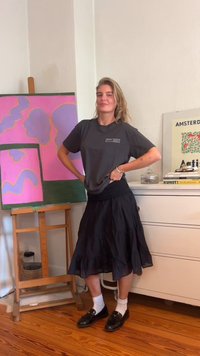 Grå T-shirt med en liten vit etikett, lager-på-lager under en svart, fyrkantig, knälång kjol. Svarta loafers med vita strumpor, trägolv.