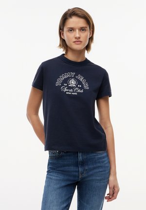 Fiatal nő, aki sötétkék Tommy Jeans Sports Club New York pólót és kék farmert visel, egyik kezét a háta mögött tartva áll.