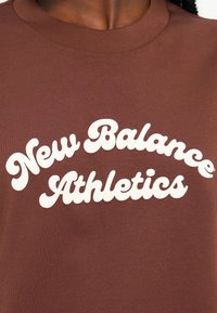 Sweatshirt castanho de gola redonda com letras brancas em negrito, estilo retro, que dizem "New Balance Athletics." Textura suave e design casual.
