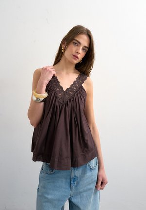 Jeune femme portant un haut sans manches marron orné de dentelle et un jean bleu, posant devant un fond blanc uni, portant de gros bracelets et des boucles d'oreilles.