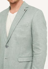 Mężczyzna w jasnozielonej marynarce na białej koszulce z dekoltem w serek, przycięty przy szyi i w połowie tułowia, stojący na jednolitym jasnym tle.