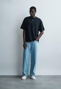 Man wearing black oversized T-shirt with "OFF" text, light blue wide-leg jeans, white sneakers, and clear glasses standing against a gray wall.