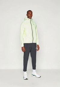 Homme se tenant debout portant une veste à capuche vert clair, un pantalon gris foncé, des chaussettes blanches et des chaussures de sport blanches sur fond uni.