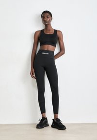 Žena stojící proti bílé zdi, oblečená v černém crop topu, černých legínách s vysokým pasem se značkou „Calvin Klein“ a černých teniskách.