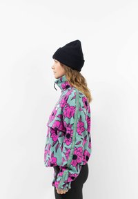Fleece pullover in teal met groot roze bloemenpatroon, hoge kraag, ritssluiting aan de voorkant en versterkte zijnaad. Gecombineerd met een zwarte gebreide beanie.
