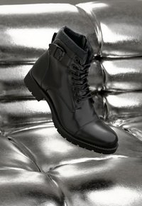 Sorte lær ankle boot med et slankt design, med snørebånd, en sidespenne og en teksturert overflate, satt mot en metallisk bakgrunn.