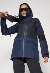 Navyblauer wasserfester Jacke mit Kapuze, die schwarze Kontrastpaneele, mehrere Reißverschlusstaschen und eine glatte Außentextur aufweist.