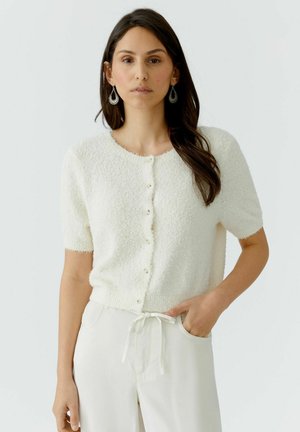 Femme aux longs cheveux bruns portant un haut blanc texturé à manches courtes avec boutons et un pantalon blanc à cordon, debout avec une main dans la poche.