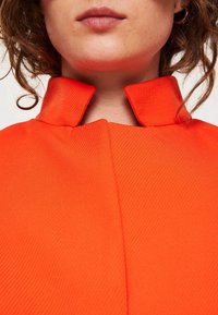 Orange ribbet stofbeklædning med en høj, struktureret krave og et sømløst design. Nærbillede, der fremhæver tekstur og farve.