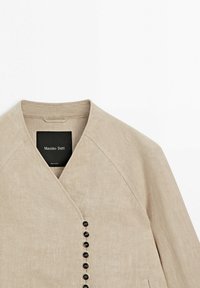 Beige linnedragt med lange ærmer, V-hals og lodret række af sorte knapper på venstre side.