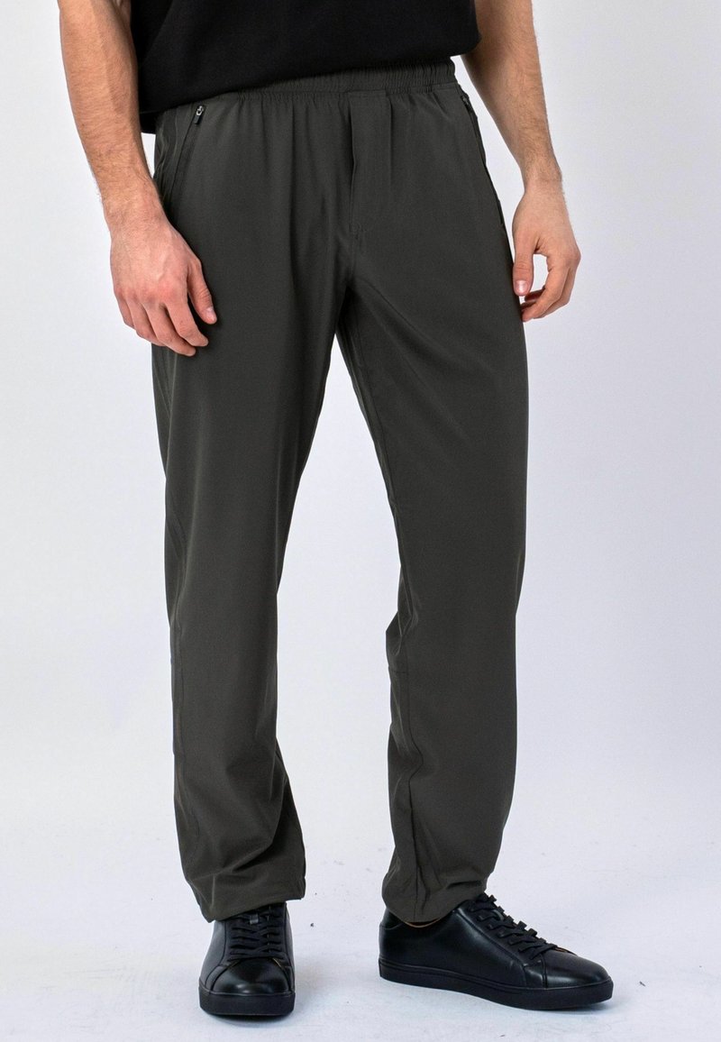 Homme portant un pantalon décontracté gris foncé avec une ceinture élastique et des baskets noires, debout devant un fond clair uni.
