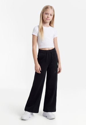 Jeune fille debout, portant un t-shirt court blanc, un pantalon de jogging noir à jambes larges avec cordon, et des baskets blanches, sur fond blanc.