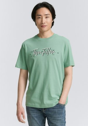 Mintgroene katoenen T-shirt met een grafische print en "Tom Tailor"-tekst in het wit. Korte mouwen en een relaxte pasvorm. Draagt met blauwe spijkerbroek.