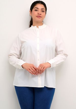 Weißes Hemd mit Rüschensaum und langen Ärmeln, aus leichtem Stoff. Kombiniert mit blauen Jeans.