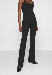 Kvinna iklädd högmidjade svarta utsvängda byxor, svart ärmlös topp och svarta spetsiga klackskor, stående mot en ljus enfärgad bakgrund.