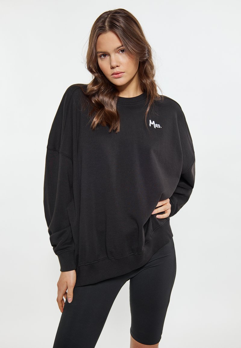 Felpa nera oversize con polsini e orlo a costine, caratterizzata da un ricamo bianco con la scritta "Mrs." sul lato sinistro del petto. Materiale in cotone morbido.