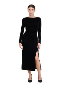 Abito lungo a maniche lunghe in velluto nero con una vestibilità elegante e uno spacco laterale. Il modello indossa tacchi neri scintillanti. Design del collo rotondo semplice.