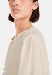 Helle beige Bluse aus transparenter Stoff, mit rundem Ausschnitt, gerafften Kanten und puffärmeln. Feine Texturdetails sind sichtbar.