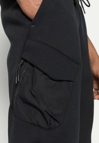 Close-up of black pants with a zipper pocket and a flap, showing part of a hand resting near the pocket.