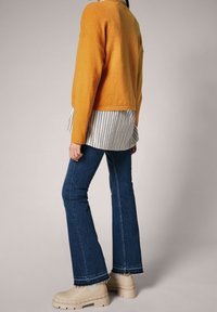 Pull orange texturé superposé sur une chemise rayée, associé à un jean évasé bleu foncé et des bottes à semelle épaisse beige.