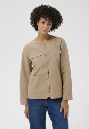 Donna con capelli ricci che indossa una giacca beige con bottoni e quattro tasche frontali, e jeans blu, in piedi davanti a uno sfondo grigio uniforme.