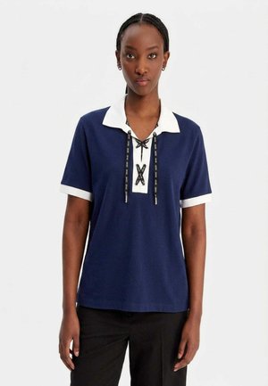 Donna che indossa una camicia blu navy a maniche corte con colletto e polsini bianchi, con dettaglio nero con lacci sul davanti, in piedi su sfondo bianco.