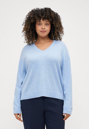 Maglione lavorato a maglia azzurro chiaro con scollo a V, motivo a treccia e polsini a costine. Indossato sopra pantaloni scuri, presenta una vestibilità comoda e una texture morbida.