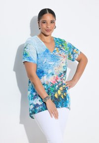 Bluzka z dekoltem w serek i krótkim rękawem w kolorze niebieskim z kwiatowym wzorem w odcieniach żółtego, różowego i zielonego. Wykonana z miękkiego materiału w luźnym kroju, zestawiona z białymi spodniami.