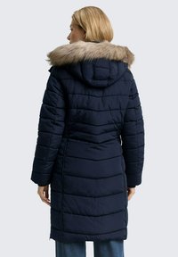 Frau in einem langen, marineblauen Steppmantel mit Kapuze, die mit einem hellbraunen Kunstfell besetzt ist, steht mit dem Rücken zur Kamera.