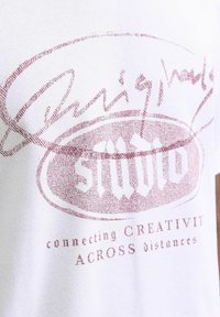 T-shirt en coton blanc avec un design graphique rouge et le texte "studio" et "connecting CREATIVIT across distances" dans une police en gras.