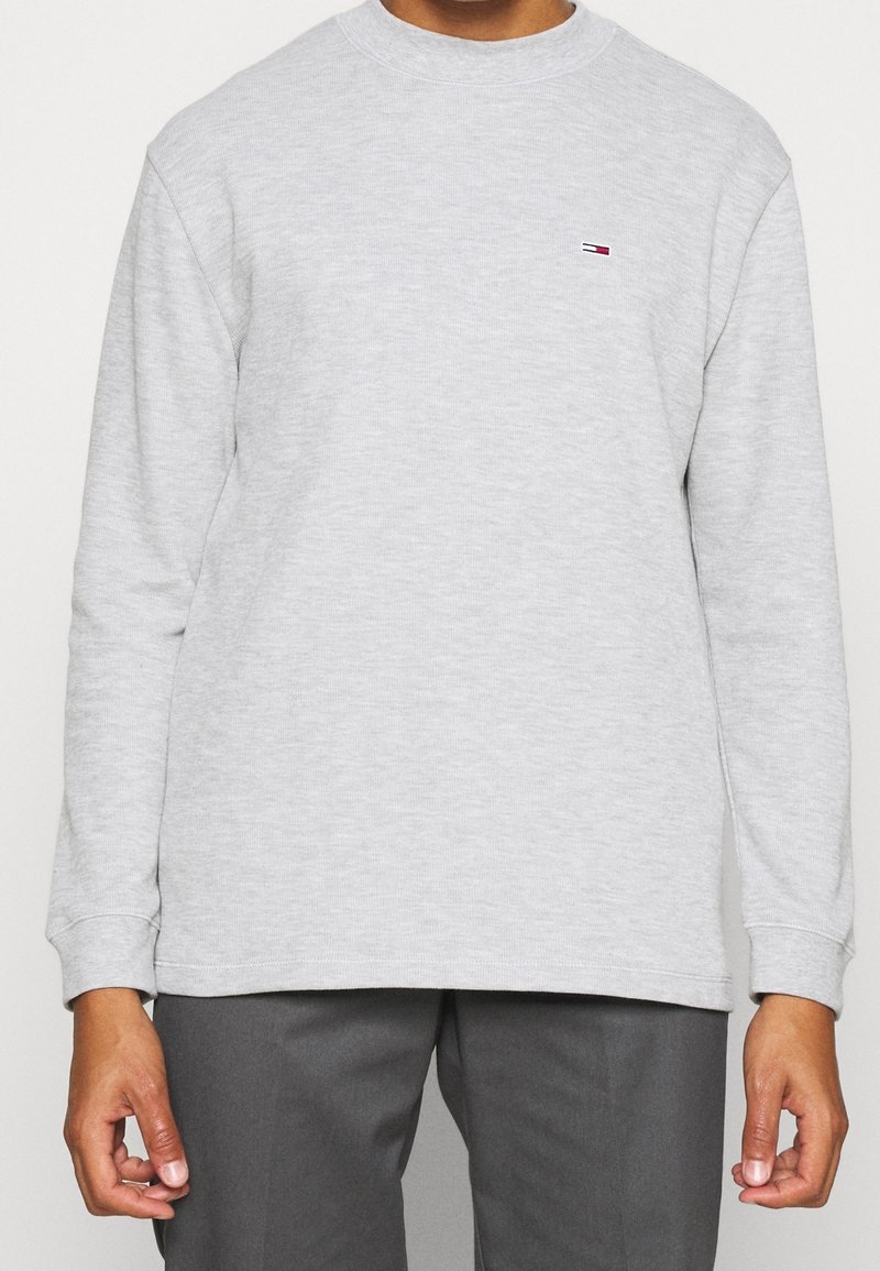 Grauer Pullover aus strukturiertem Stoff, mit einem gerippten Rundhalsausschnitt und langen Ärmeln. Kleines Logo-Detail auf der Brust in Rot und Blau.