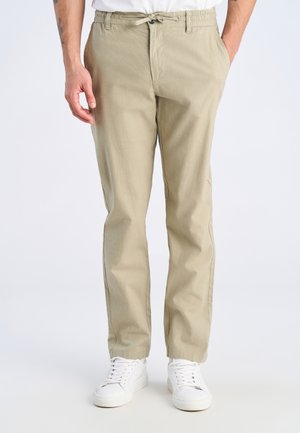 Uomo che indossa pantaloni casual beige con vita elasticizzata e coulisse, abbinati a sneaker bianche, in piedi davanti a uno sfondo semplice.
