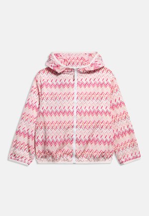 Børnejakke med hætte i lyserød og rød zigzag-mønster, hvid lynlås foran, lange ærmer og elastisk kant.
