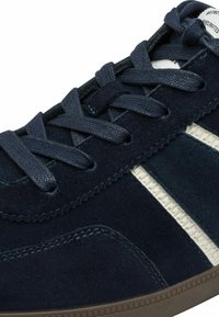 Primo piano di una sneaker in suede blu navy con lacci abbinati, striscia laterale bianca testurizzata e bordo della suola in gomma marrone.