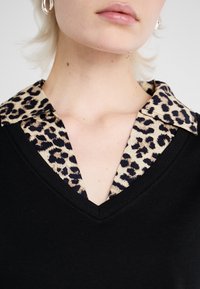 Čierny top s výstrihom do V a golierom z leopardieho vzoru. Golier má hnedé a čierne škvrny, ktoré kontrastujú so solidnou čiernou látkou. Hladká textúra.