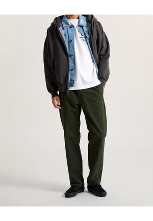 Mann trägt einen geschichteten Freizeitlook mit schwarzem Hoodie, blauer Jeansjacke, weißem bedrucktem T-Shirt, olivgrüner Hose und schwarzen Sneakers.