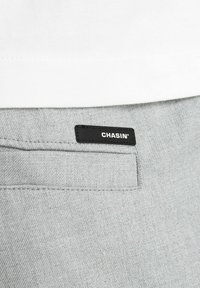Tissu gris avec une poche cousue et une étiquette noire indiquant "CHASIN'" au-dessus, sous un vêtement blanc rentré.