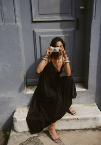Black maxi dress with a flowing silhouette, worn with silver jewelry. The individual holds a camera, sitting on stone steps against a gray wall.