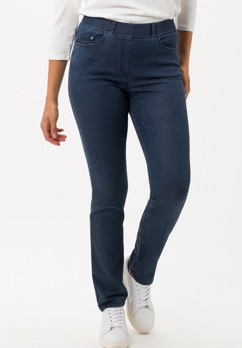 Jeans in denim di colore blu scuro con design aderente, dotati di tasche frontali e una texture liscia. Abbinati a sneaker bianche per creare contrasto.